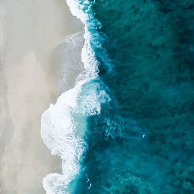 An aerial view of a wave breaking onto pale sand from deep turquoise water.