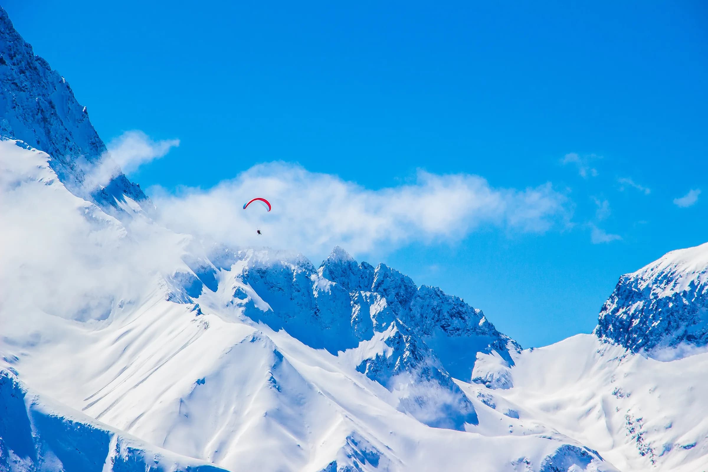 A paraglider flying over snow-covered peaks under a clear blue sky.