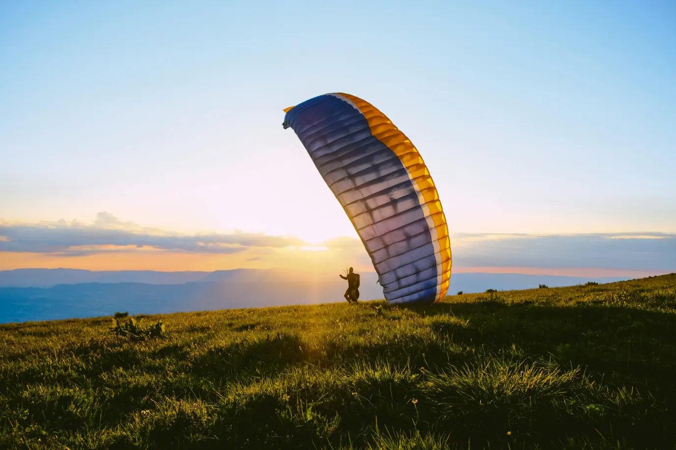 A paraglider standing on a grassy hilltop at sunset, the canopy inflated and towering beside them, the low sun behind.