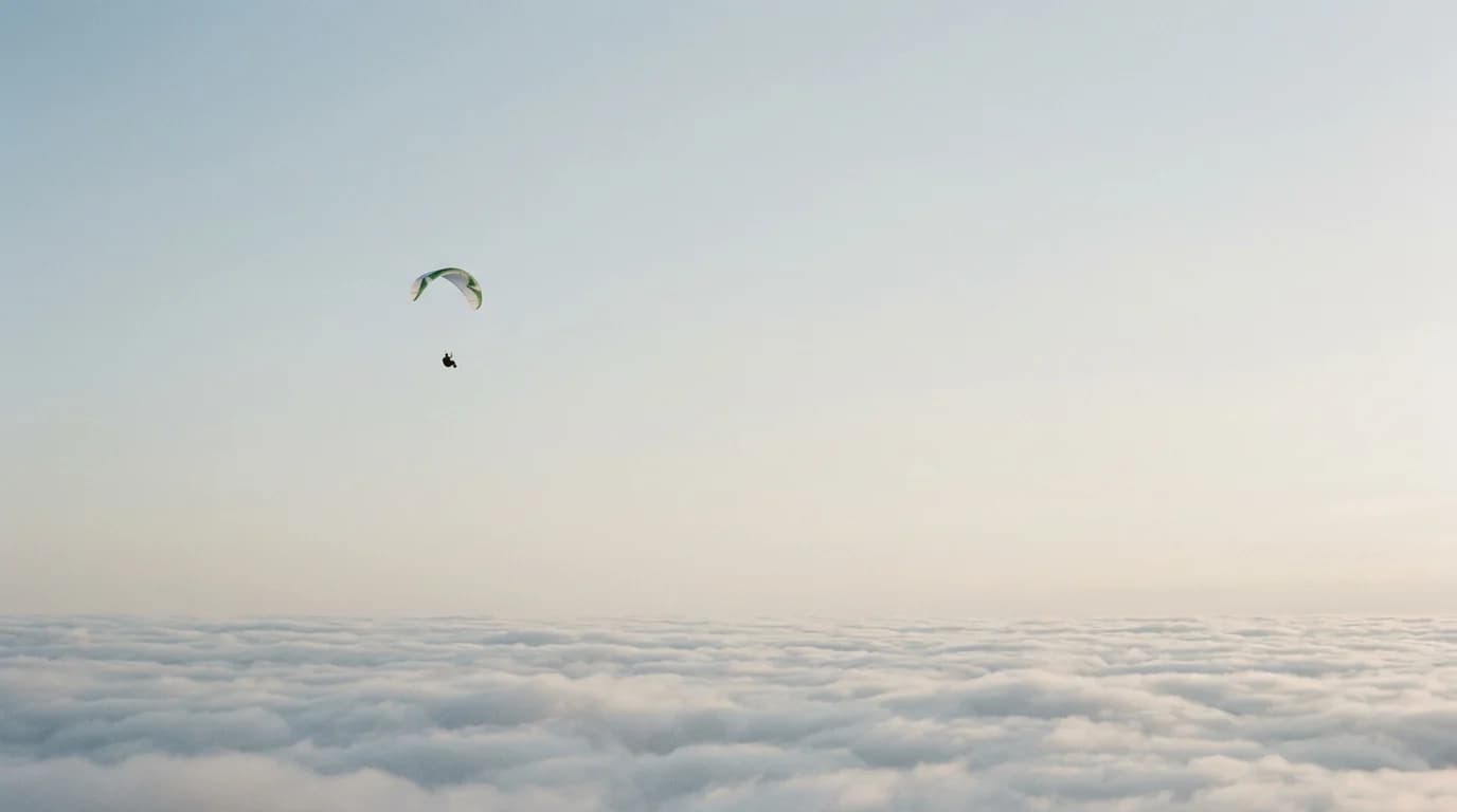 A lone paraglider high in a wide, empty, pale-blue sky.